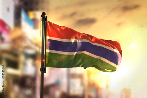 The Gambia Flag Against City Blurred Background At Sunrise Backlight photo