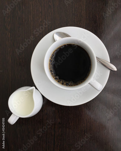 top view of black coffee in white cup with mini milk jug on black wooden table.