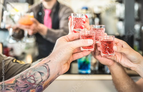 Group of drunk friends toasting cocktails at bar restautant - Food and beverage concept on nightlife moments - Defocused bartender serving drinks on background - Focus on hands cheering red shot glass