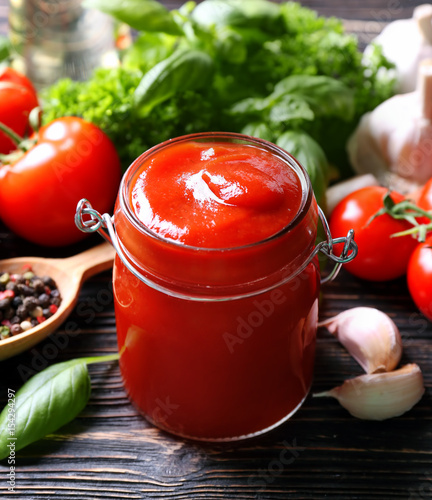 Composition of ketchup in jar and ingredients on wooden background, closeup