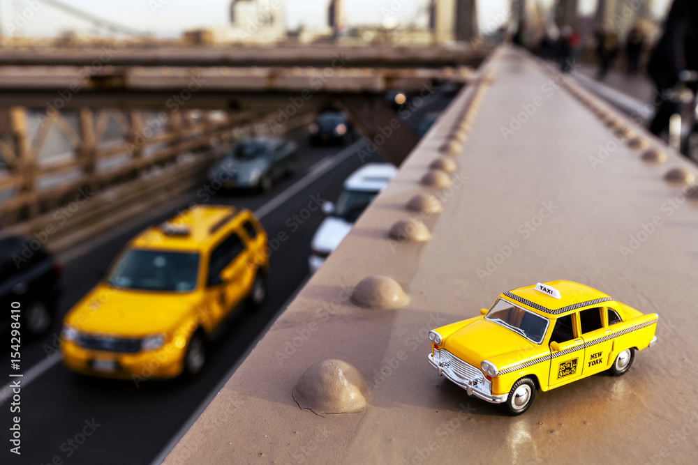 Model of a classic yellow taxi on a steel beam on Brooklyn Bridge, with a real taxi passing on the lower level asphalt road.