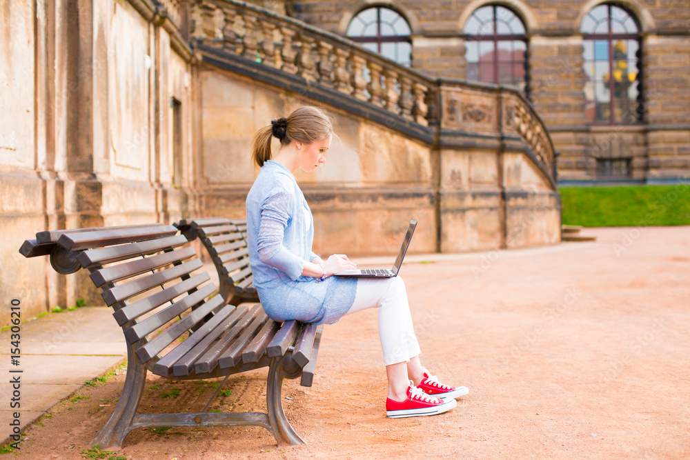 A girl is sitting on a bench and working on a laptop in a European city A girl is sitting on a bench and working on a laptop in a European city
