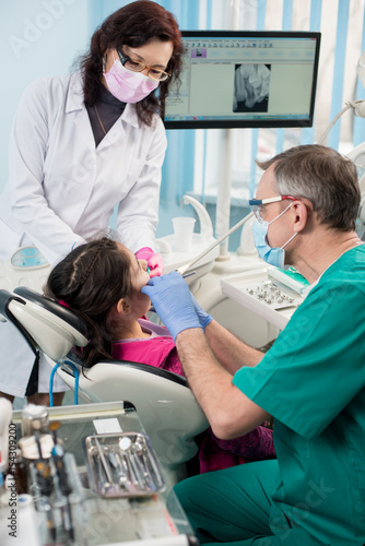 Young girl with on the first dental visit. Senior pediatric dentist with nurse treating patient teeth at the dental office. On the background screen with X-ray the patient s teeth. Dental equipment