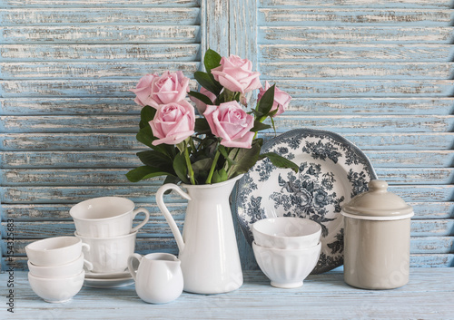 Pink roses in a white enameled pitcher, vintage crockery on blue wooden rustic background. Kitchen still life in vintage style. Flat lay