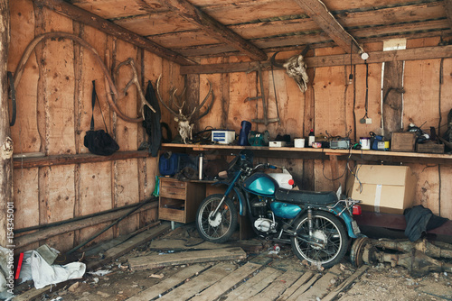 Old Blue-Green Motorbike In Picturesque Barn.Vintage Motorcycle In Old Hangar Against A Wall With Deer Antlers, A Bison Head And Many Interesting Rare Objects. Old Barn With Old Moped And Wooden Walls photo