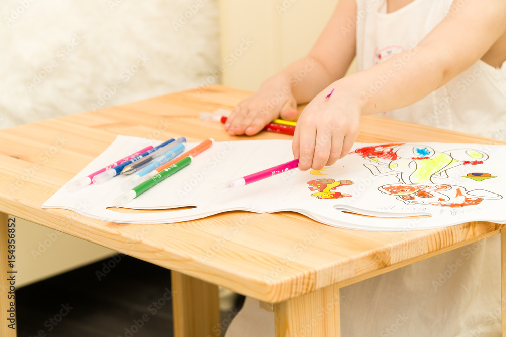 Active little preschool age child, cute toddler girl with blonde curly hair, drawing picture on paper using colorful pencils and felt-tip pens, sitting at wooden table indoors at home or kindergarten Active little preschool age child, cute toddler girl with blonde curly hair, drawing picture on paper using colorful pencils and felt-tip pens, sitting at wooden table indoors at home or kindergarten
