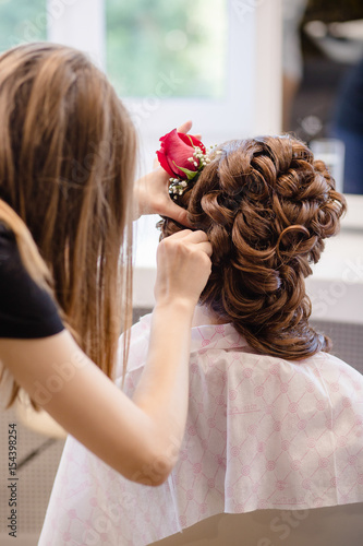 Female hairdresser standing and making hairstyle to cute lovely young woman in beauty salon. Hairstyle with red roses.