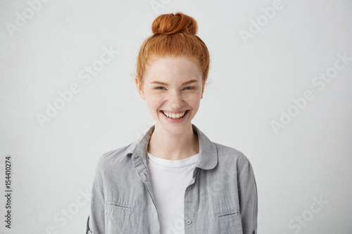 Beautiful ginger freckled girl with mysterious smile posing indoors at blank grey studio wall. Pretty woman with hair bun smiling broadly, showing white straight teeth, enjoying leisure time at home