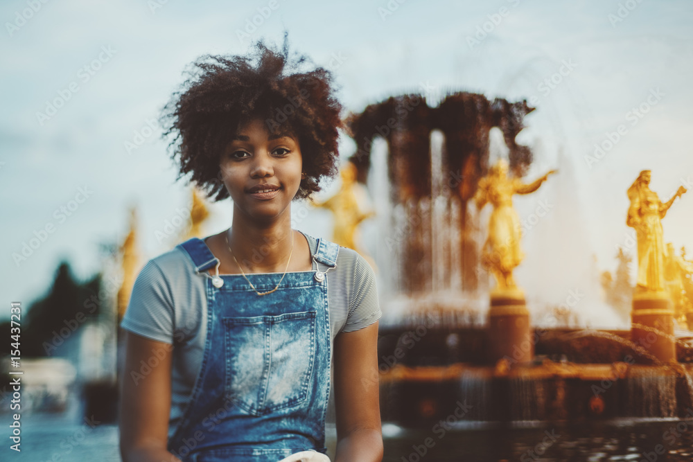 Tilt-shift portrait of young cute afro american girl in jeans overalls  sitting in front of beautiful fountain "Friendship of Nations" in Russia,  Moscow with golden statues and pool on sunny evening Stock