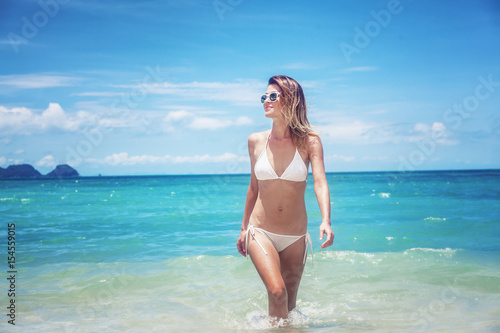 Beautiful young woman in a white bikini walking along a tropical beach with azure water and white sand © olezzo