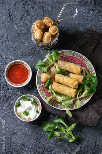 Fried spring rolls with red and white sauces, served in traditional china plate and fry basket with fresh green salad over black texture background. Flat lay, space. Asian food