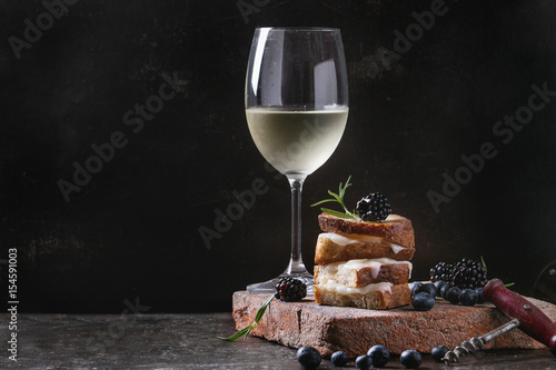 Grilled sandwich with melted goat cheese, blackberry, blueberry, rosemary and honey, served on terracotta board with glass of cold white wine. over dark background. Summer appetizer.