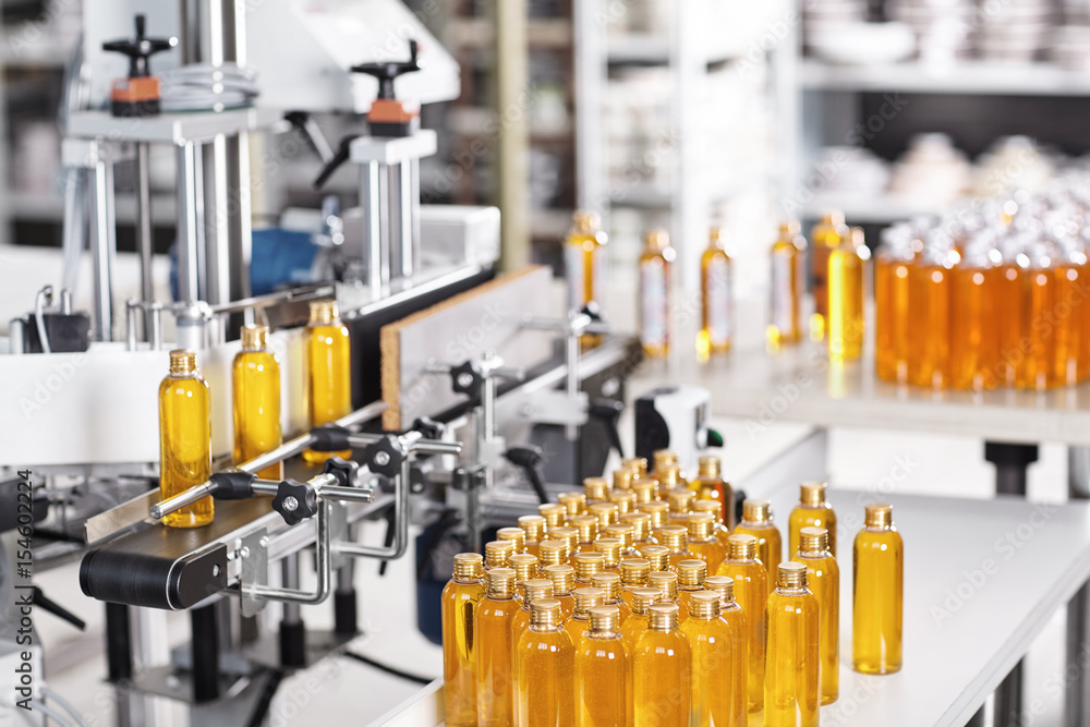 Bottles filled with yellow thick substance standing on desk and conveyor line. Production line of cosmetics products on big factory.Automated process of creation shampoo on factory.Research concept Bottles filled with yellow thick substance standing on desk and conveyor line. Production line of cosmetics products on big factory.Automated process of creation shampoo on factory.Research concept