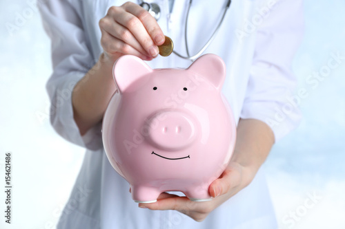 Female doctor putting coin into piggy bank, close up. Concept of medical insurance