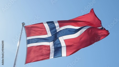Norwegian flag blowing towards right on a pole, towards clear blue sky in gale wind, with heavy gusts 