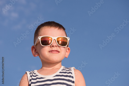 cheerful little boy wearing sunglasses and striped shirt on blue sky background