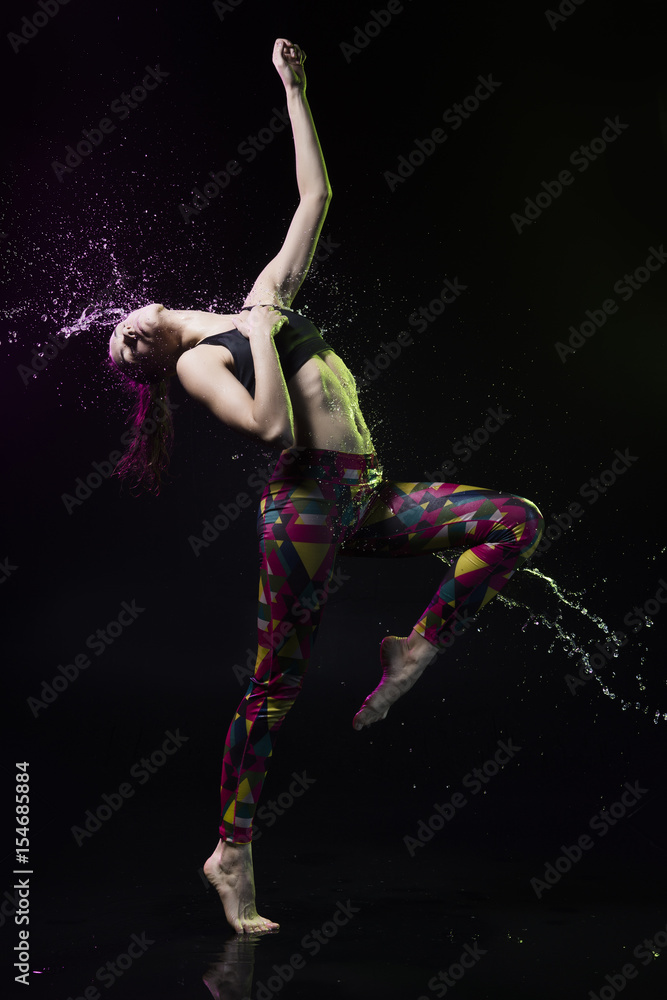 The girl dances on the floor covered with water on a black background and water splashes around her