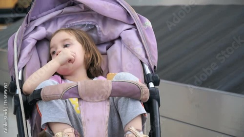 Portrait of a child sitting in a stroller on the background of a tape for luggage. One lost suitcase passes by the girl. Girl 4-5 years of European appearance, flew by plane to the resort on vacation.