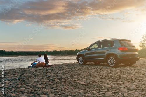 Portrait of happy young adult couple with dog on roadtrip. Man sitting on plaid with woman and watching sunset near river. Outdoor picnic concept.