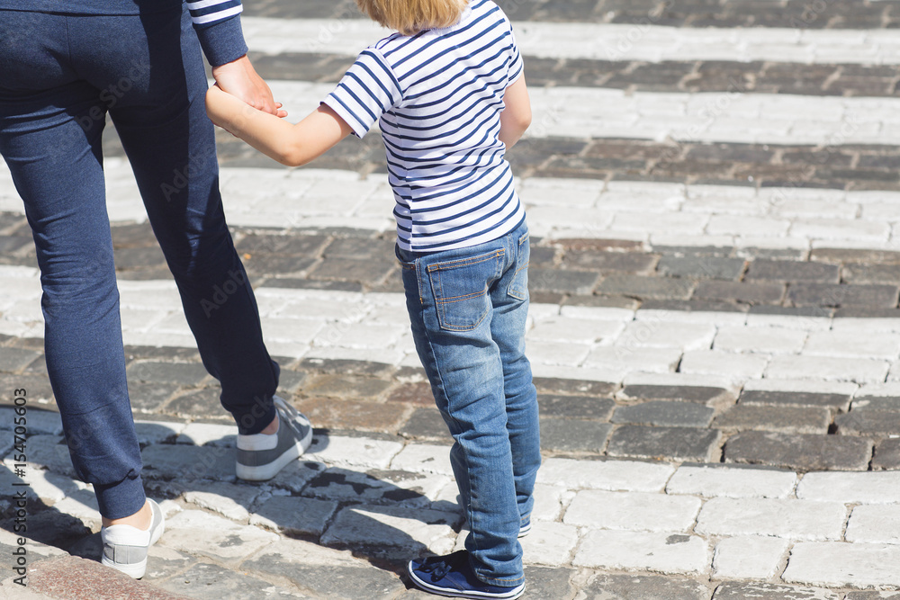 Young mother and her little child crossing the street holding by the hand. Boy and adult woman walking on the crosswalk on the road with hard traffic.