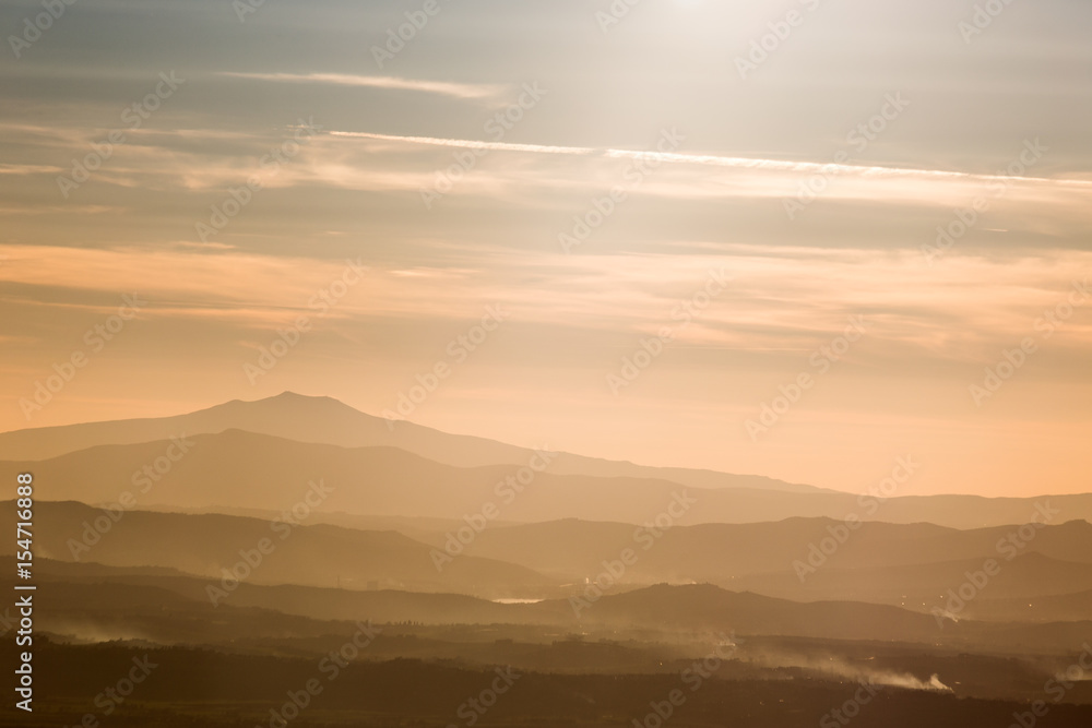 Various layers of mountains and hills in the middle of a golden, orange light during sunset, with clouds also reflecting light, and smoke on the right bottom corner