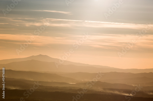 Various layers of mountains and hills in the middle of a golden, orange light during sunset, with clouds also reflecting light, and smoke on the right bottom corner