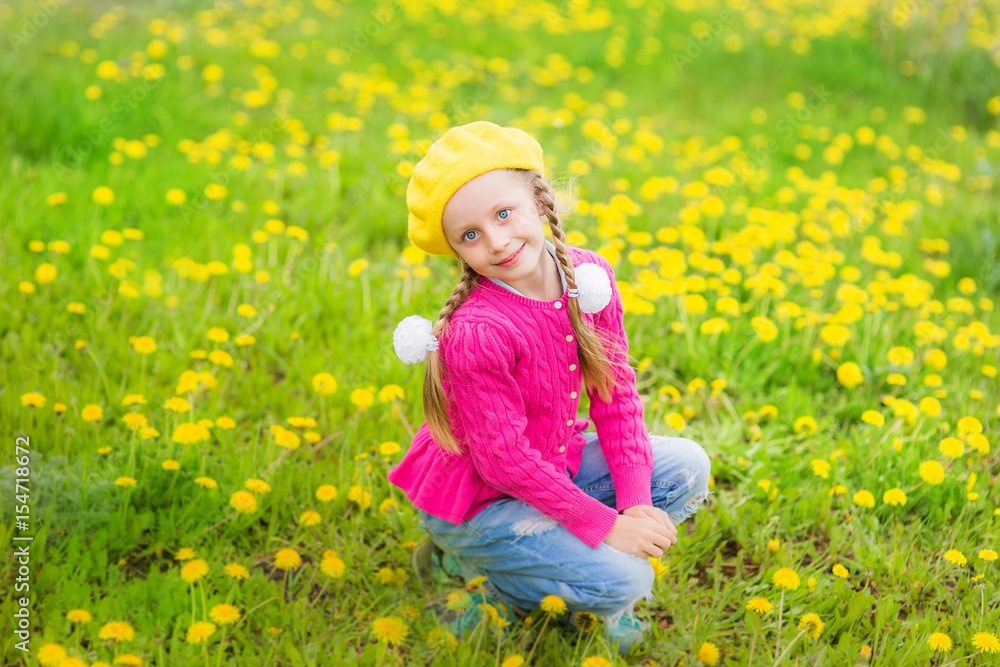 Cute beautiful girl in a yellow hat with two pigtails sits in a field of yellow dandelions Cute beautiful girl in a yellow hat with two pigtails sits in a field of yellow dandelions