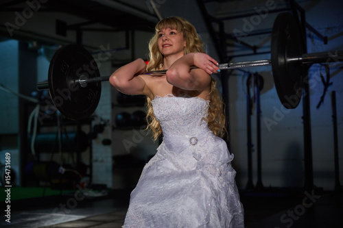 Beautiful and muscular bride blonde in a wedding dress doing an exercise with a barbell in a gym, crossfit photo