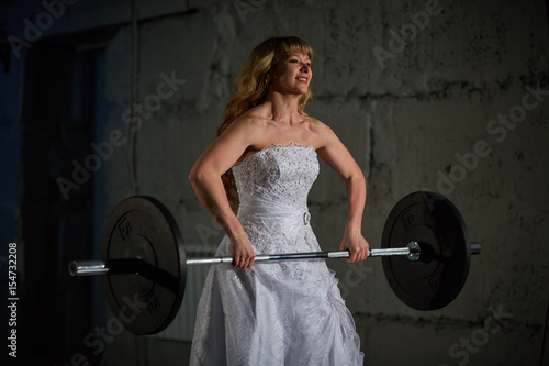 Beautiful and muscular bride blonde in a wedding dress doing an exercise with a barbell in a gym, crossfit