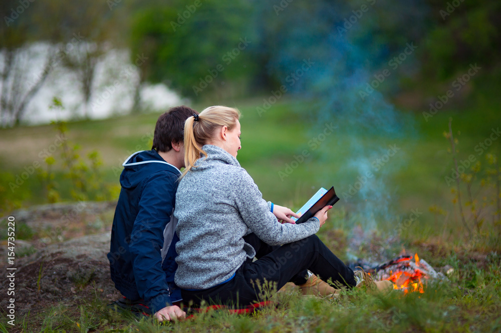 Young couple hikers looking at map. A Man and woman sitting near the fire embracing. Travel, vacation, holidays and adventure concept. Forest Mountain landscape background