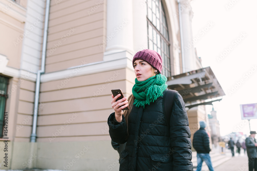 Fototapeta premium The girl on the street during a winter time is very puzzled with something. On her a lilac cap, a green scarf and a blue jacket. The girl holds phone in hand.