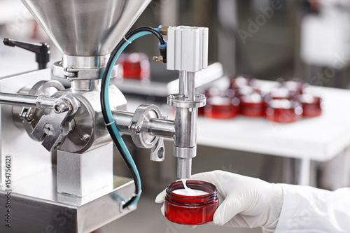 Hard mechanism of packing ready beauty cream into red container. A factory worker dressed in white lab gown and gloves holding red container under tap pouring ready cream. Production process of cream