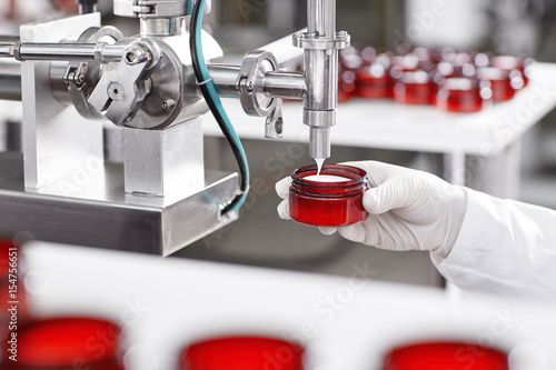Portrait of big factory process of creating lotion for skincare. Researcher holding container under big metal tube pouring beauty cream isolated over working process at factory and many containers
