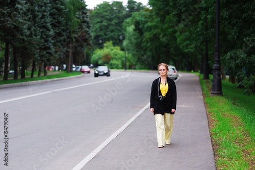 Attractive middle-aged woman in sunglasses with a camera around his neck calmly walking down side of the highway on blurred background moving cars and green trees.