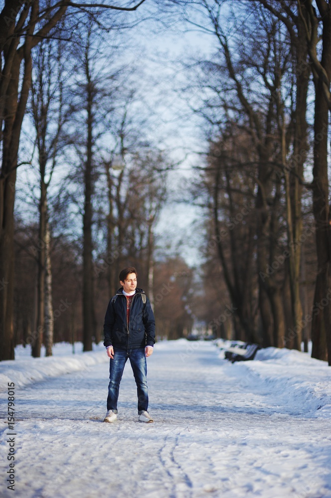 Young serious man in jacket and jeans, no hat, posing in winter city Park on a snowy sidewalk among tall trees.