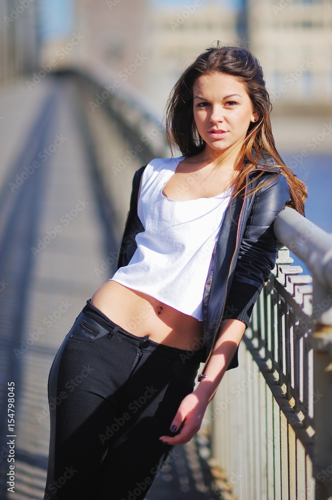 Portrait of an cute woman, dressed in a short white t-shirt, black leather jacket and trousers at hips. She stands leaning against railing of bridge, in centre of Saint-Petersburg on a Sunny day