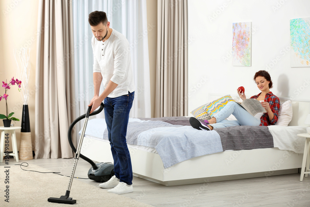 Woman reading newspaper on bed while man hoovering floor at home Woman reading newspaper on bed while man hoovering floor at home