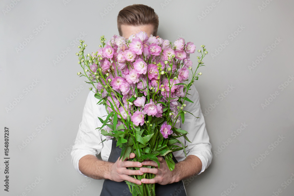Young florist hiding behind beautiful bouquet on grey background