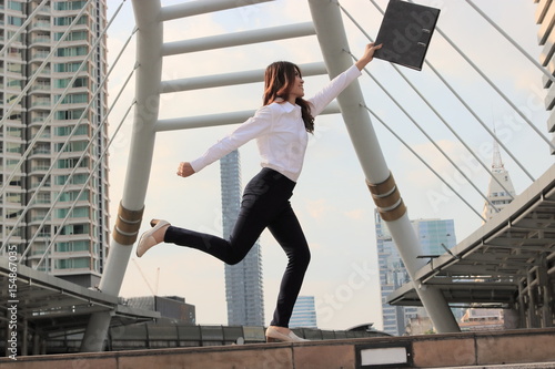 Successful young Asian business woman raising her arms with document file at cityscape background.