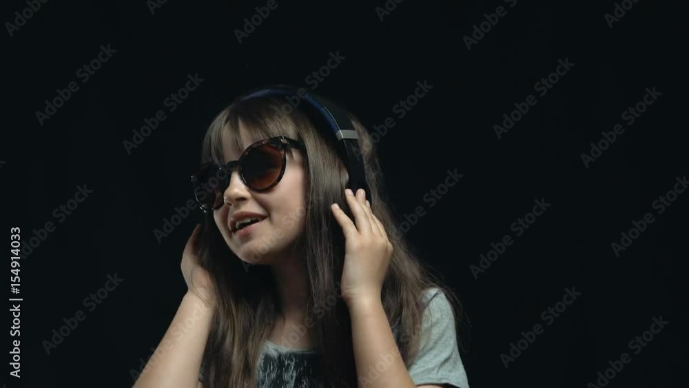 Studio shot of cute teenager girl wears headphones and sunglasses before black background, listening rhytmical music, youth freedom concept