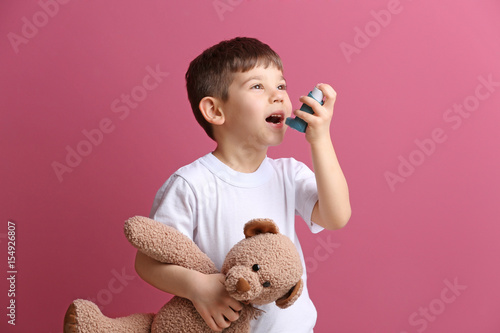 Cute little boy holding inhaler and toy bear on color background. Allergy concept photo
