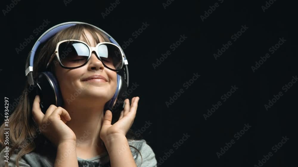 Studio shot of cute teenager girl wears headphones and sunglasses before black background, listening rhytmical music, youth freedom concept Studio shot of cute teenager girl wears headphones and sunglasses before black background, listening rhytmical music, youth freedom concept