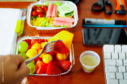 Healthy man using a fork to eat in the office a delicious fresh fruit salad, and a blurred mediterranean-Style fresh salad with hamon on aluminium box on wooden table, above view photo