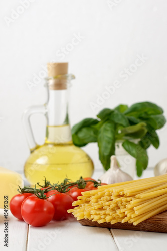 Culinary still life with dry pasta bucatini, fresh tomatoes and basil, bottle of oil on light background.