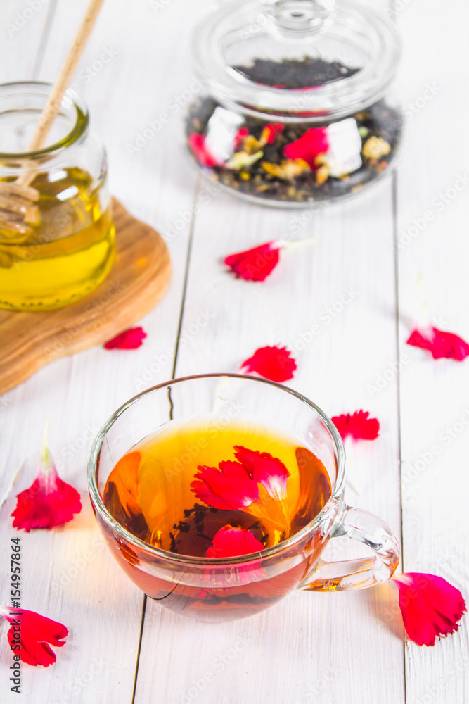 A cup of tea, a can of honey and a jar with a black herbal floral tea on a white wooden table. Top view. A cup of tea, a can of honey and a jar with a black herbal floral tea on a white wooden table. Top view.