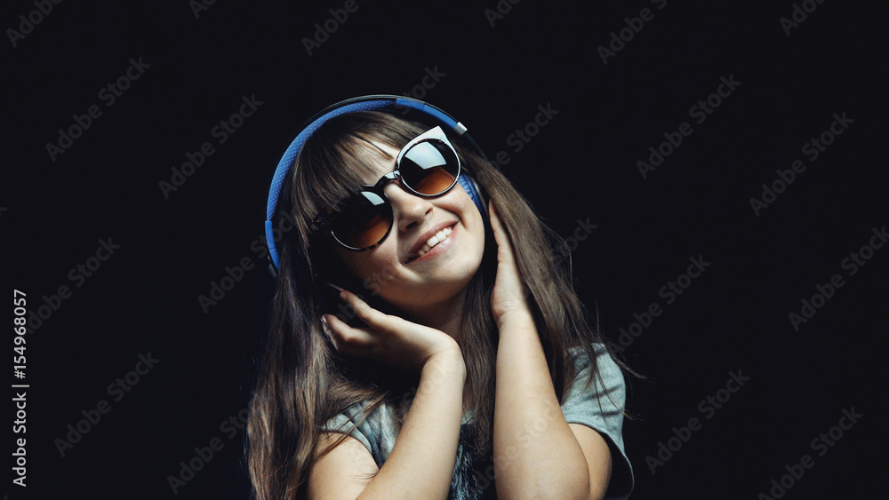 Studio shot of cute teenager girl wears headphones and sunglasses before black background, listening rhytmical music, youth freedom concept