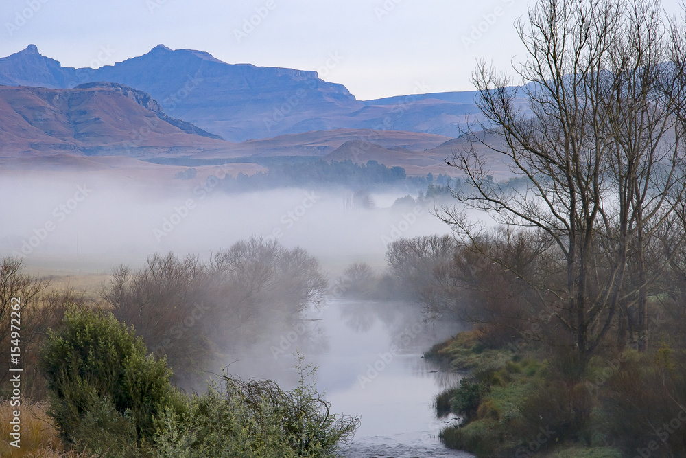 A river runs through the foothills of the Drakensberg Mountain Range at Underberg in South Africa.  This landscape-orientation takes in the mountains and a section of the Ngangwane River in the mist.