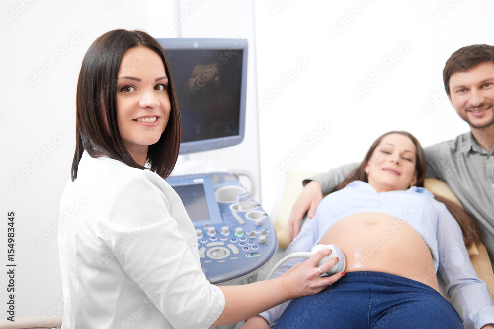 Fototapeta premium Husband embracing his pregnant wife by and carrying, when female doctor using ultrasound equipment diagnosing woman. Smiling doctor at modern hospital with patients making diagnostic procedure.