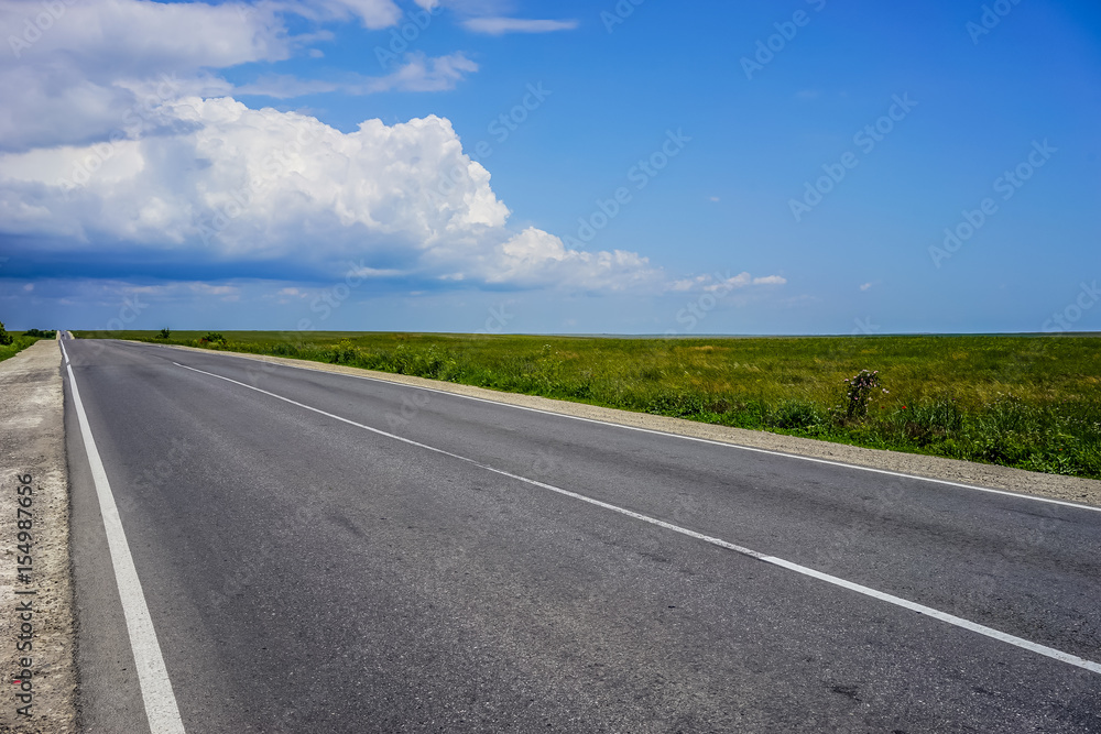 A long highway with no cars on the overgrown grass of the steppe under a blue cloudy sky