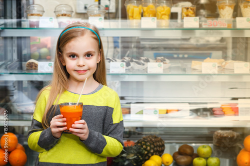 A little girl stands near a showcase with glass of juice in a cafe